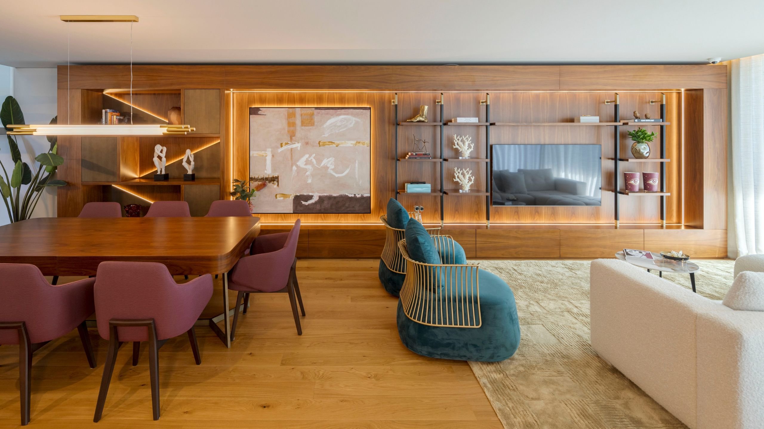 Vista ampla de sala de estar moderna e sala de jantar em open space, destacando o sofá claro, poltronas azuis, mesa para sala de jantar em madeira e móvel de TV com estantes integradas.