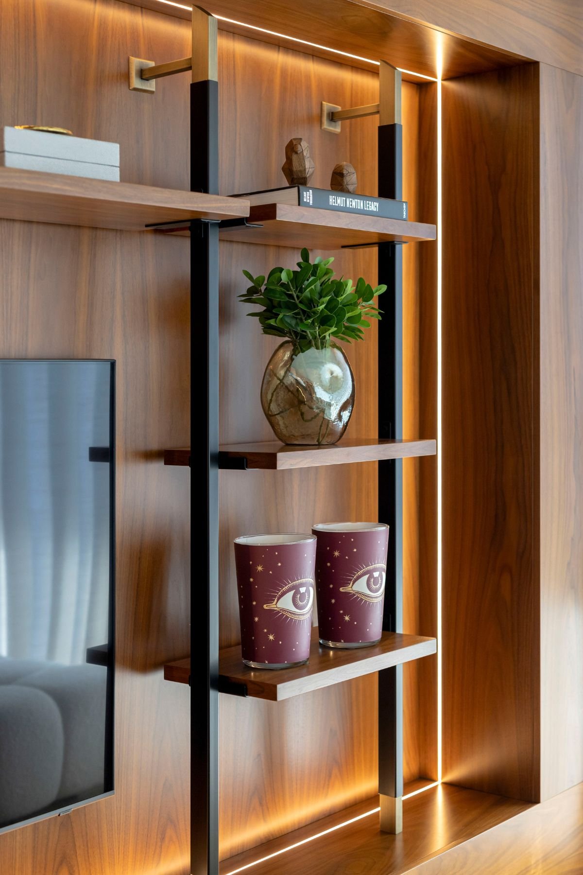 Pormenor de estante metálica sobre painel de madeira no móvel TV, decorada com vasos decorativos, elementos de inspiração marinha, livro e iluminação quente.