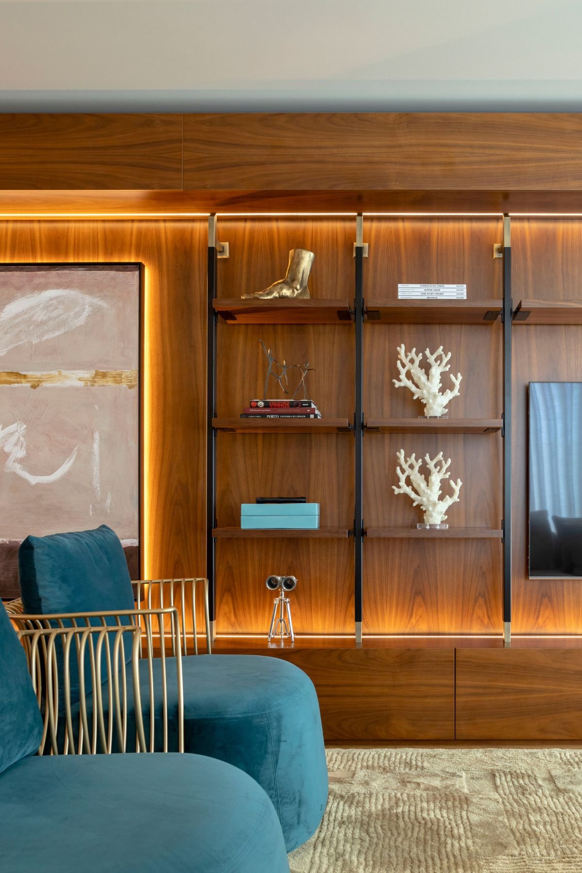 Pormenor da estante metálica com prateleiras em madeira, peças decorativas em tom coral e dourado, ao lado da poltrona azul e do painel iluminado com quadro decorativo.
