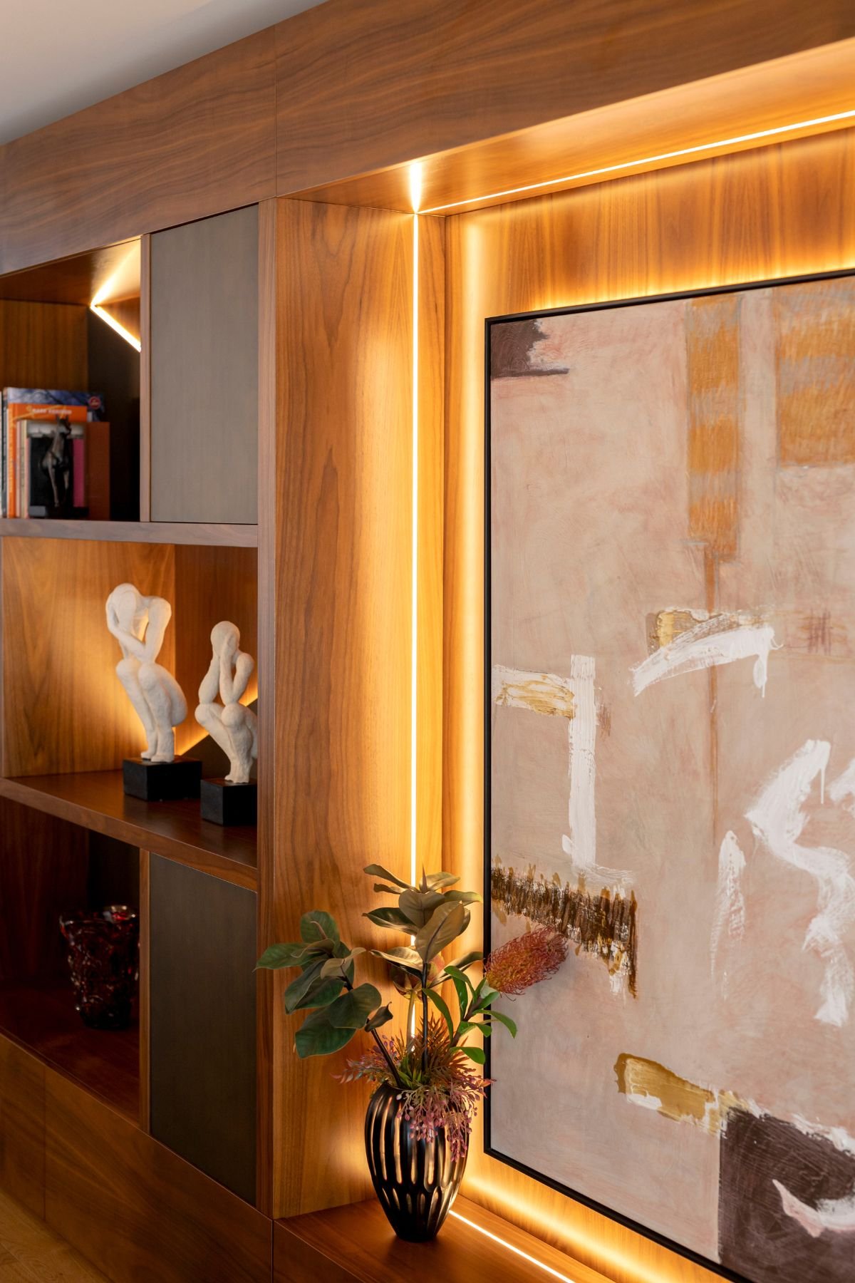 Pormenor da iluminação LED embutida no móvel de madeira da sala, realçando quadro decorativo abstrato, vaso com plantas e estatuetas decorativas.