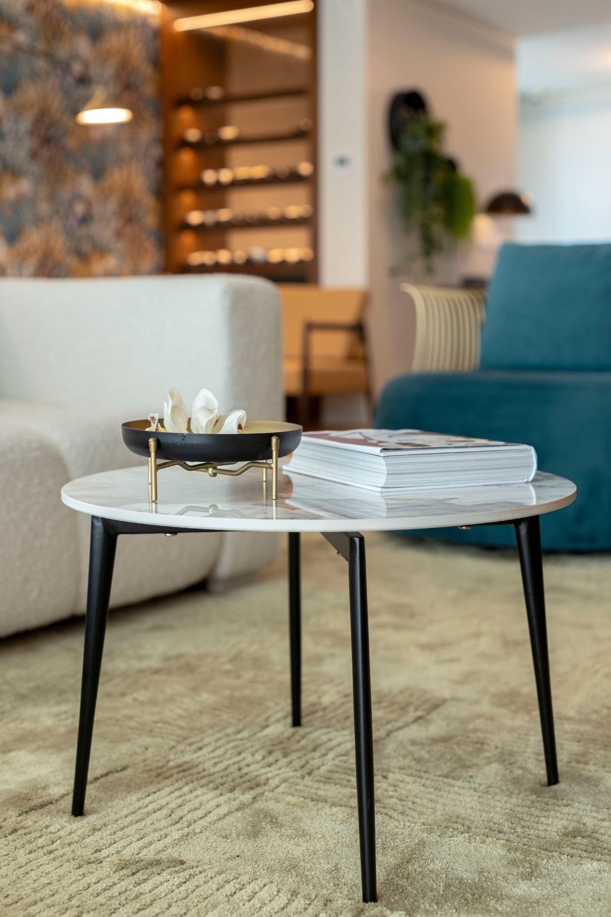 Plano detalhado de mesa de centro redonda em mármore com pés pretos, livro e centro de mesa decorativo, sob tapete de sala com o sofá e a poltrona ao fundo.