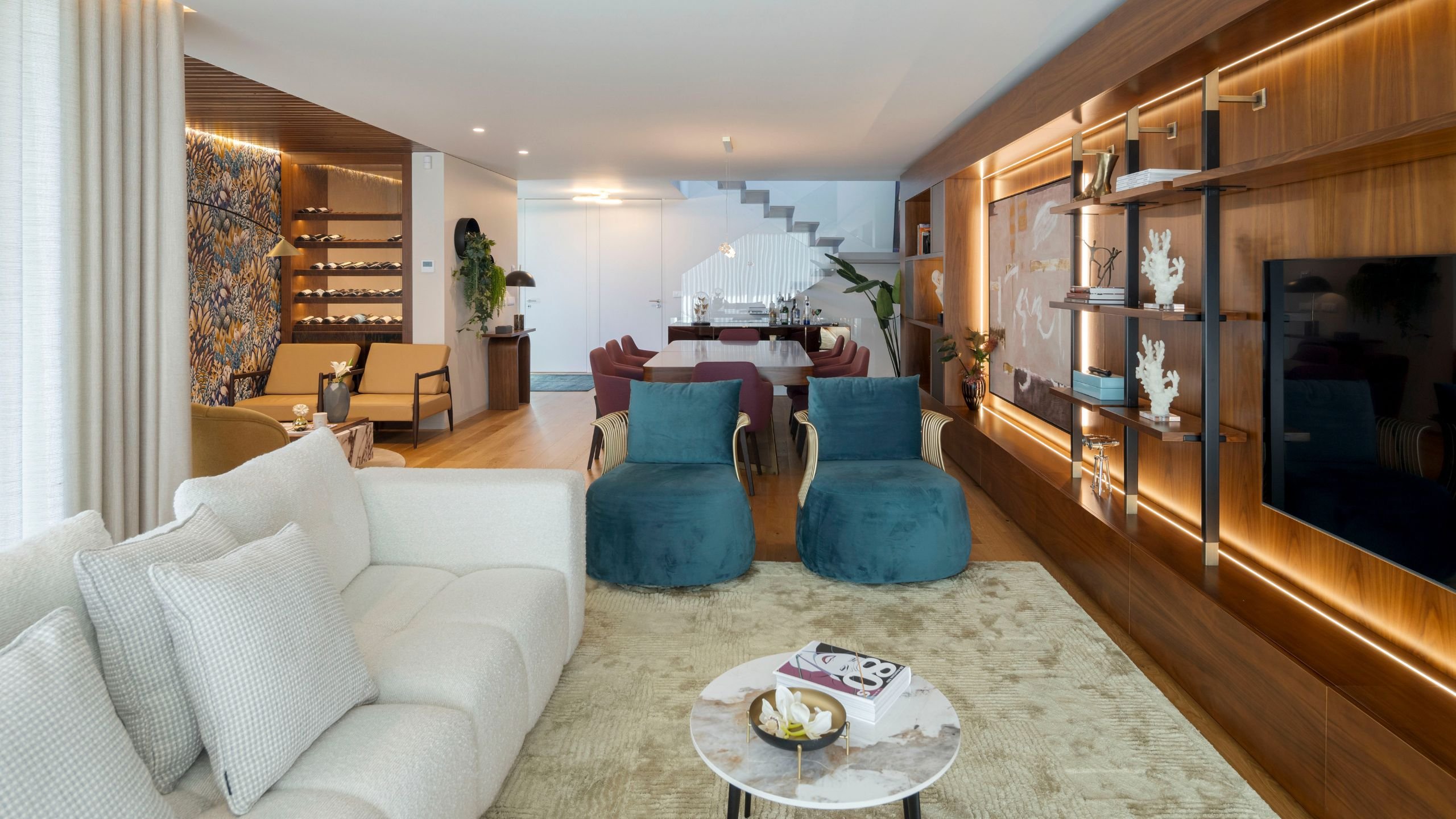 Vista geral da sala de estar e sala de jantar em open space de casa moderna, com sofá Antónia, poltronas azuis, mesa de centro e móvel de TV em madeira iluminado.