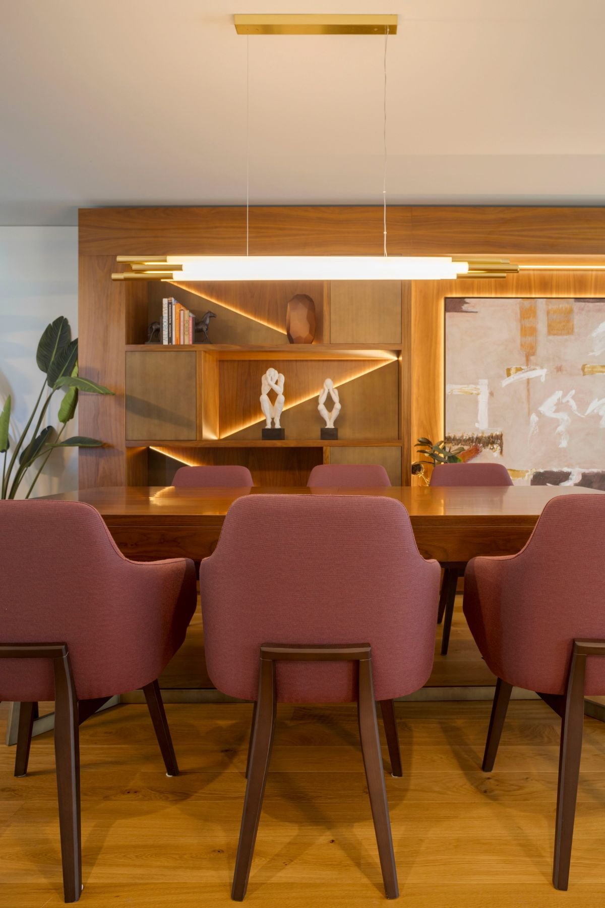 Vista focada das cadeiras estofadas em tom vinho na mesa de jantar em madeira, com candeeiro de teto tubular suspenso e móvel iluminado ao fundo.
