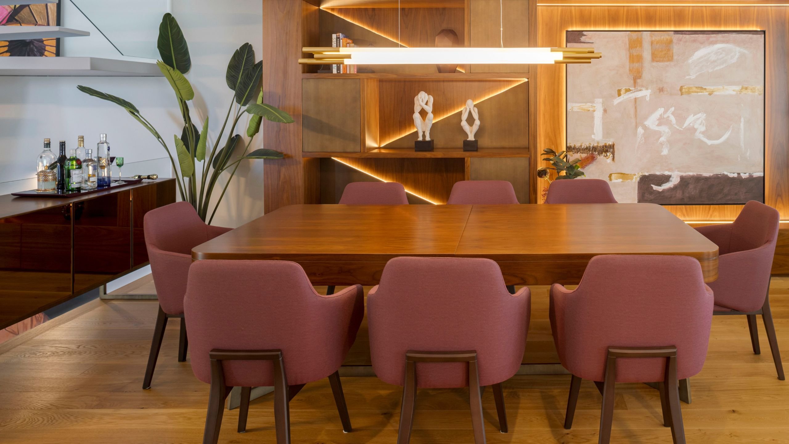 Sala de jantar moderna em open space com mesa de jantar rectangular em madeira para oito lugares, cadeiras cor de vinho, aparador com bar e iluminação suspensa.
