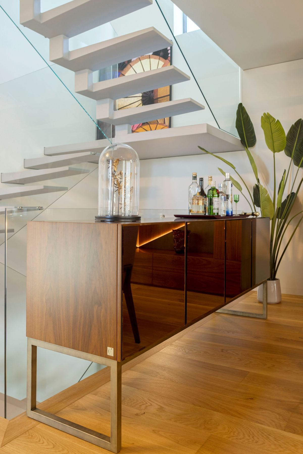 Aparador de sala moderno com portas espelhadas a funcionar como bar de apoio, posicionado junto a escadas interiores com guarda-corpo em vidro.