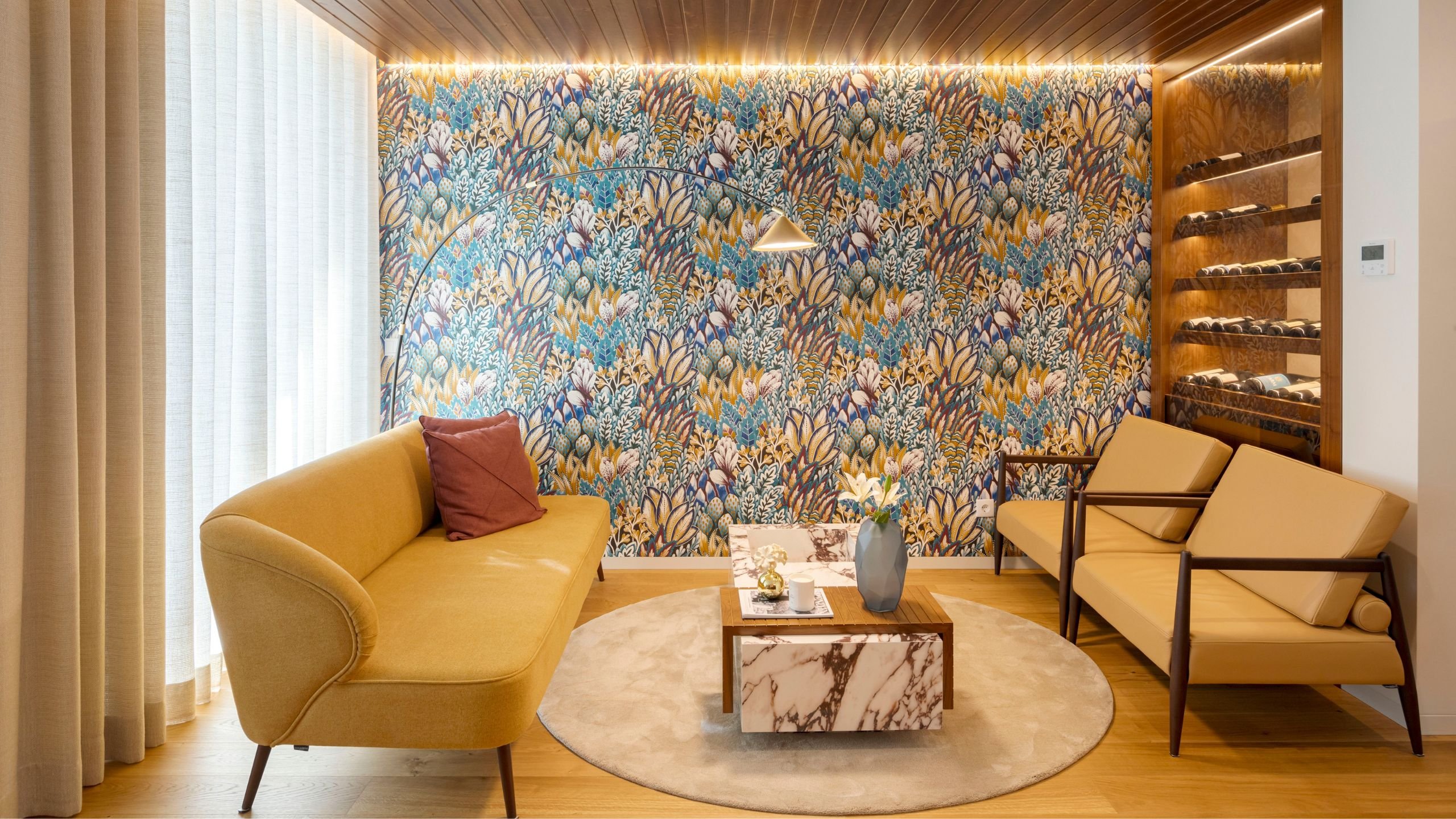Canto de lounge aconchegante com papel de parede floral colorido, sofá amarelo torrado, dois cadeirões e mesa de centro em mármore.