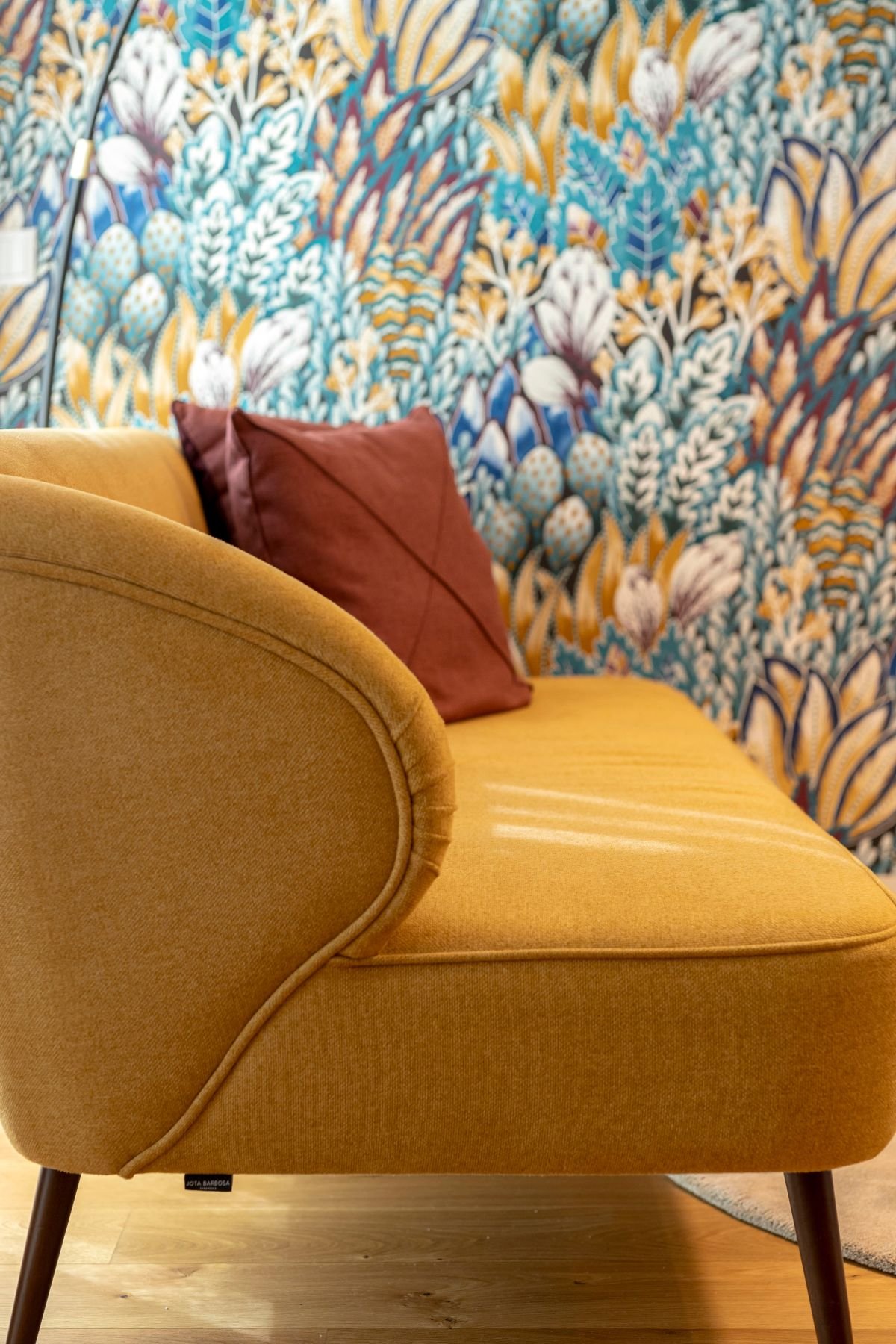Detalhe do estofo do sofá amarelo torrado com almofada em tom vinho sobre o fundo de papel de parede com padrão de folhas azuis e amarelas.