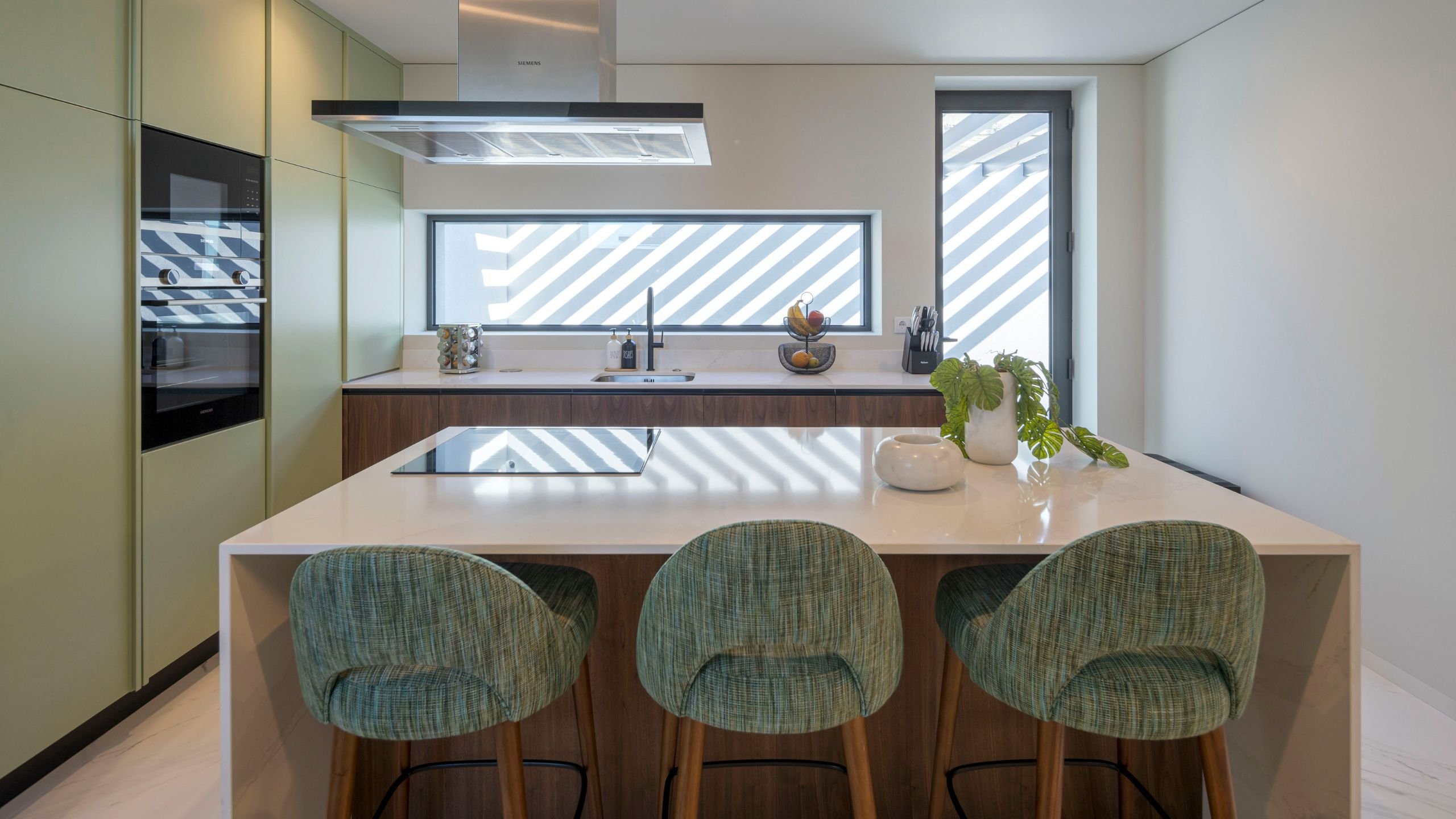 Cozinha moderna com ilha central em bancada branca, três bancos altos com estofo verde e pés de madeira, armários em verde-alface e eletrodomésticos encastrados.