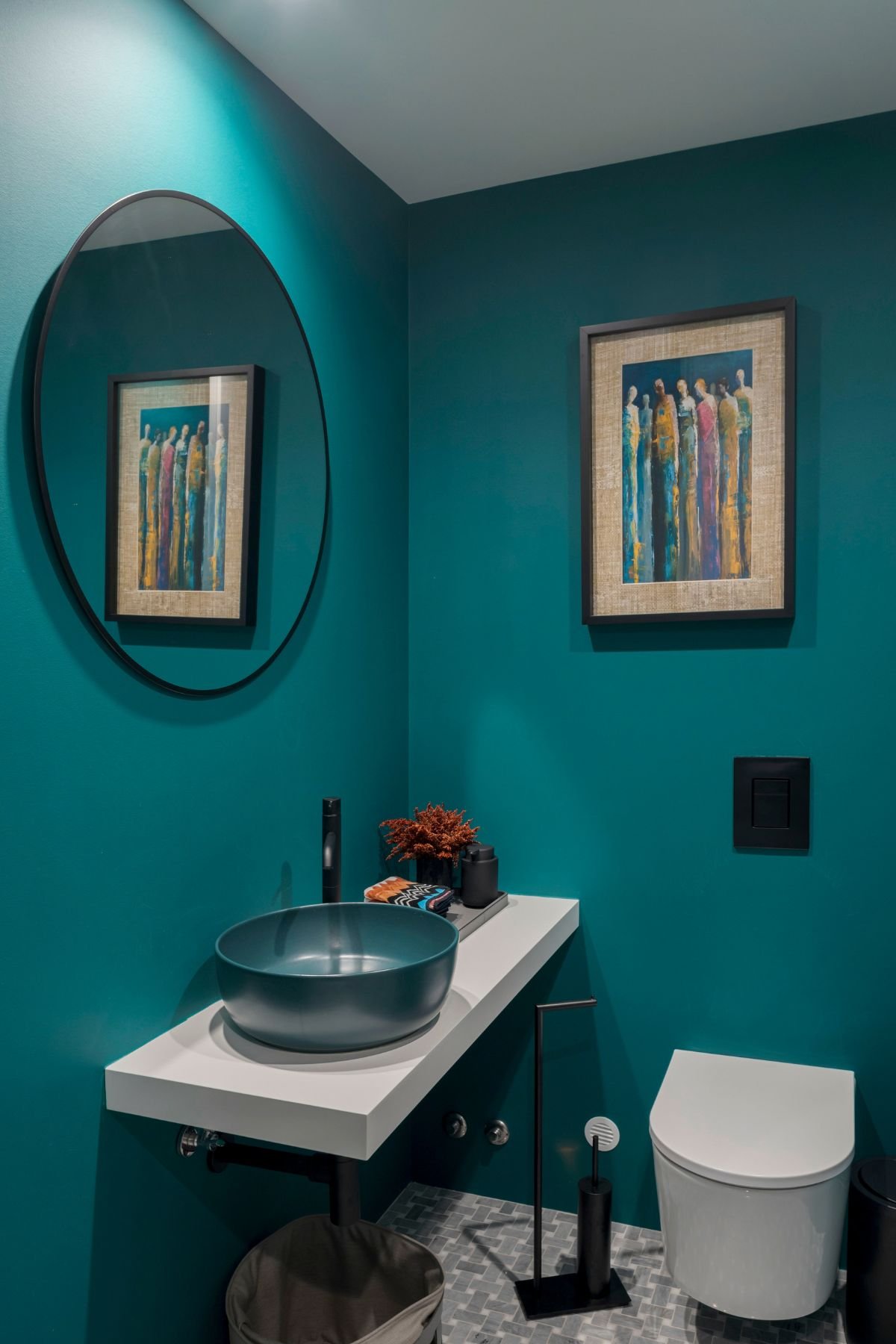 Casa de banho social com paredes azul-petróleo, lavatório de pousar redondo azul sob bancada suspensa branca, espelho redondo e quadro decorativo.