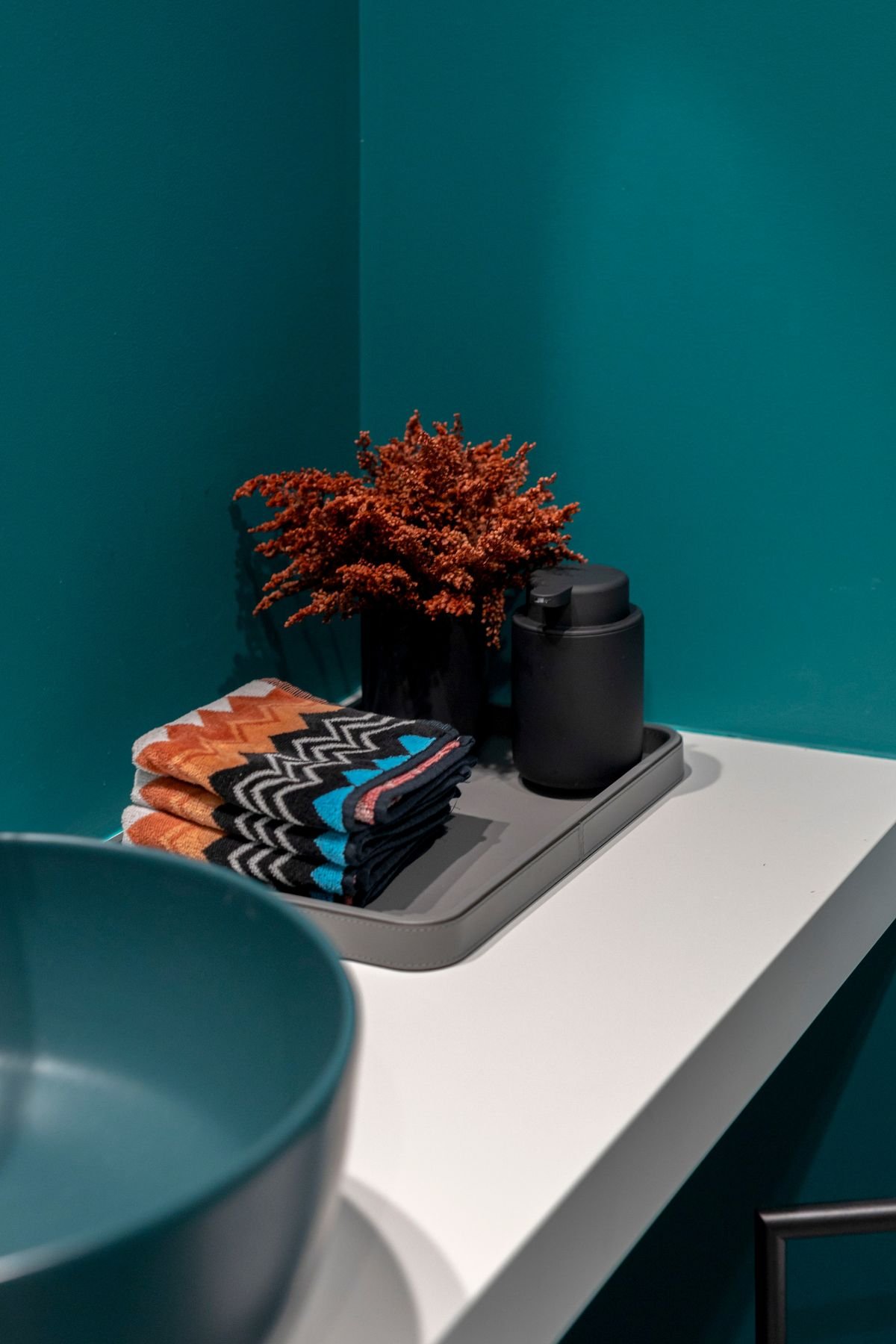 Detalhe da bancada de casa de banho branca com lavatório azul, tabuleiro cinzento com doseador preto, planta decorativa terracota e toalhas dobradas.