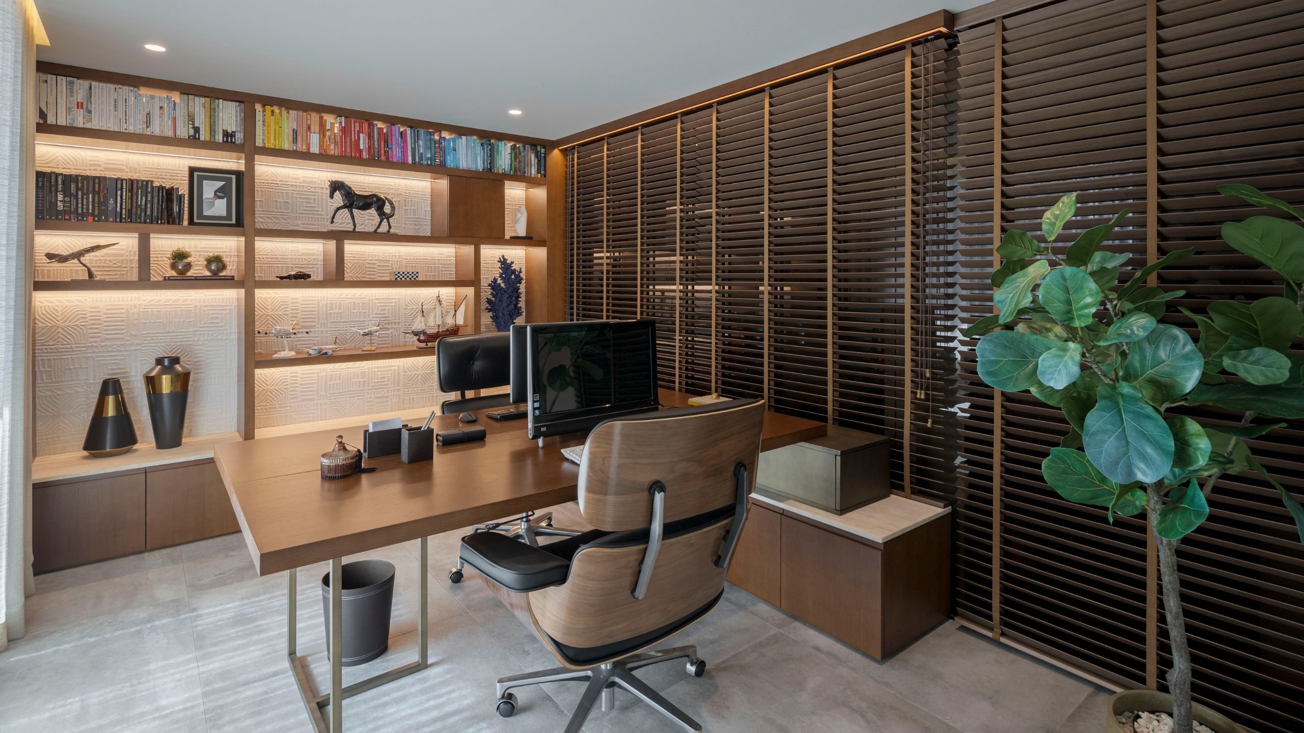 Escritório de trabalho em casa com secretária em madeira, cadeiras de escritório pretas, estante iluminada com livros e persianas venezianas em madeira.