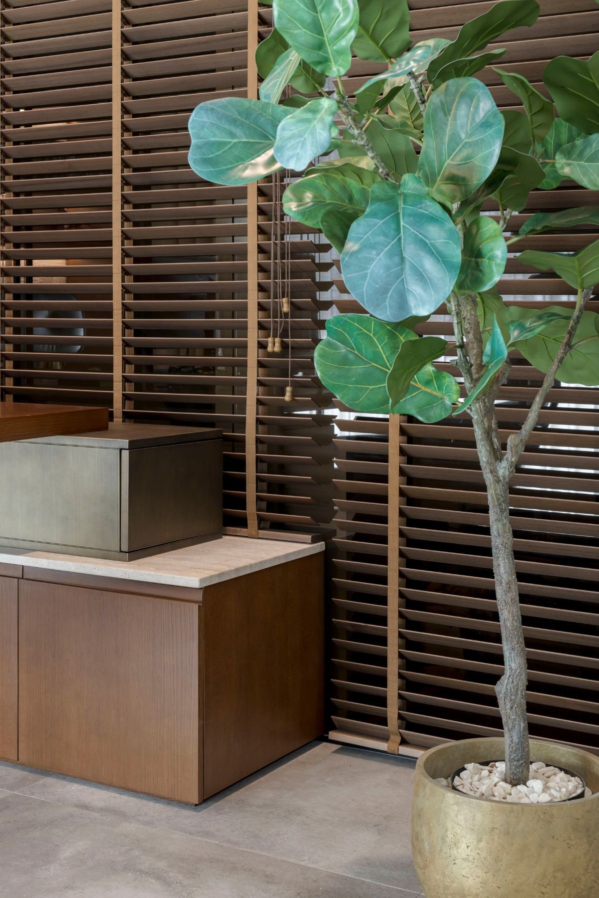 Pormenor do canto do escritório com móvel de apoio em madeira, persianas venezianas e planta de interior decorativa em vaso.