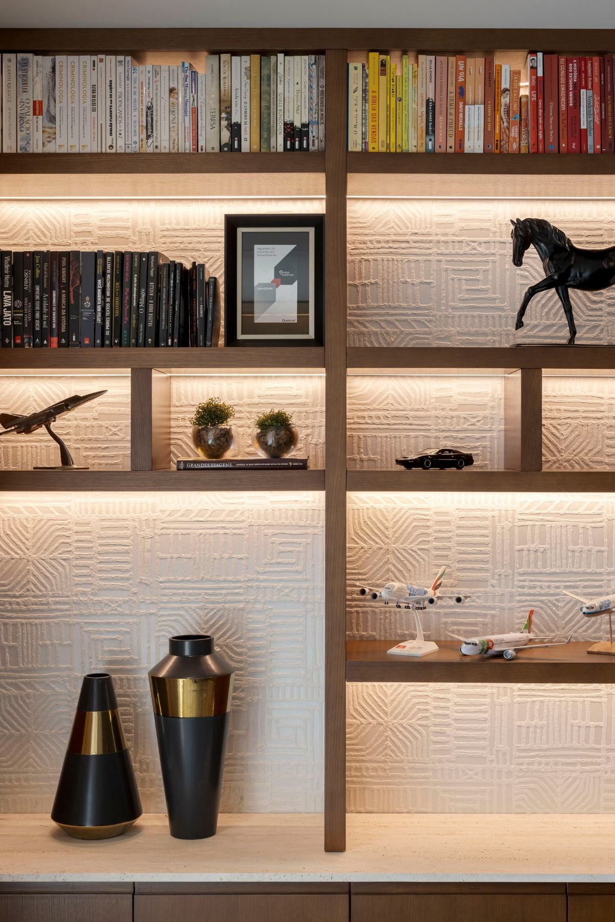 Vista focada na estante de madeira iluminada com LED, destacando o papel de parede texturizado branco, livros, maquetes de aviões e figuras decorativas.