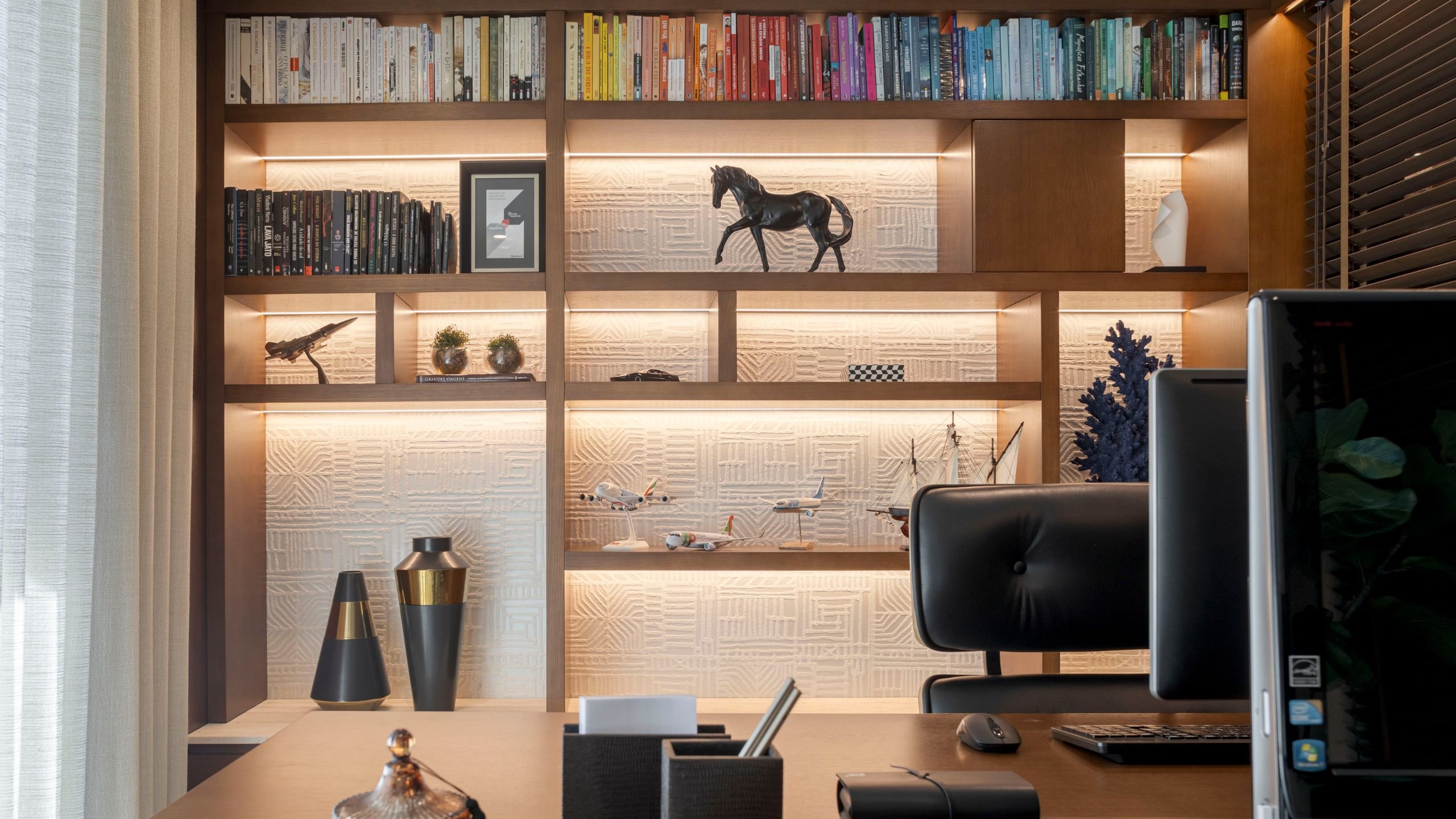 Estante de escritório por medida em madeira com iluminação embutida e fundo texturizado branco, com vista parcial do posto de trabalho e monitor.