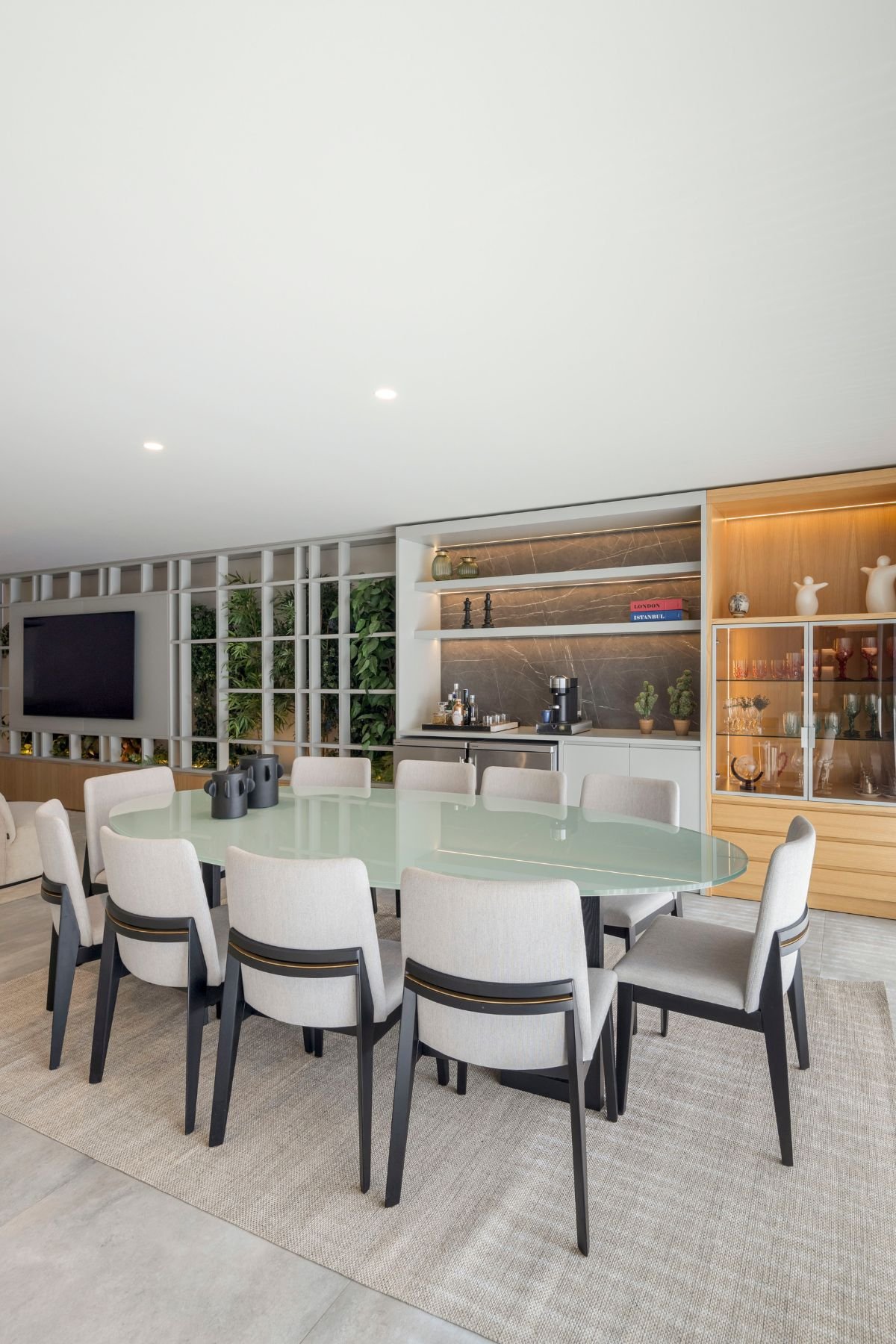 Zona de jantar integrada com mesa oval de tampo em vidro, cadeiras modernas estofadas a cinzento e móvel bar com vitrina iluminada.