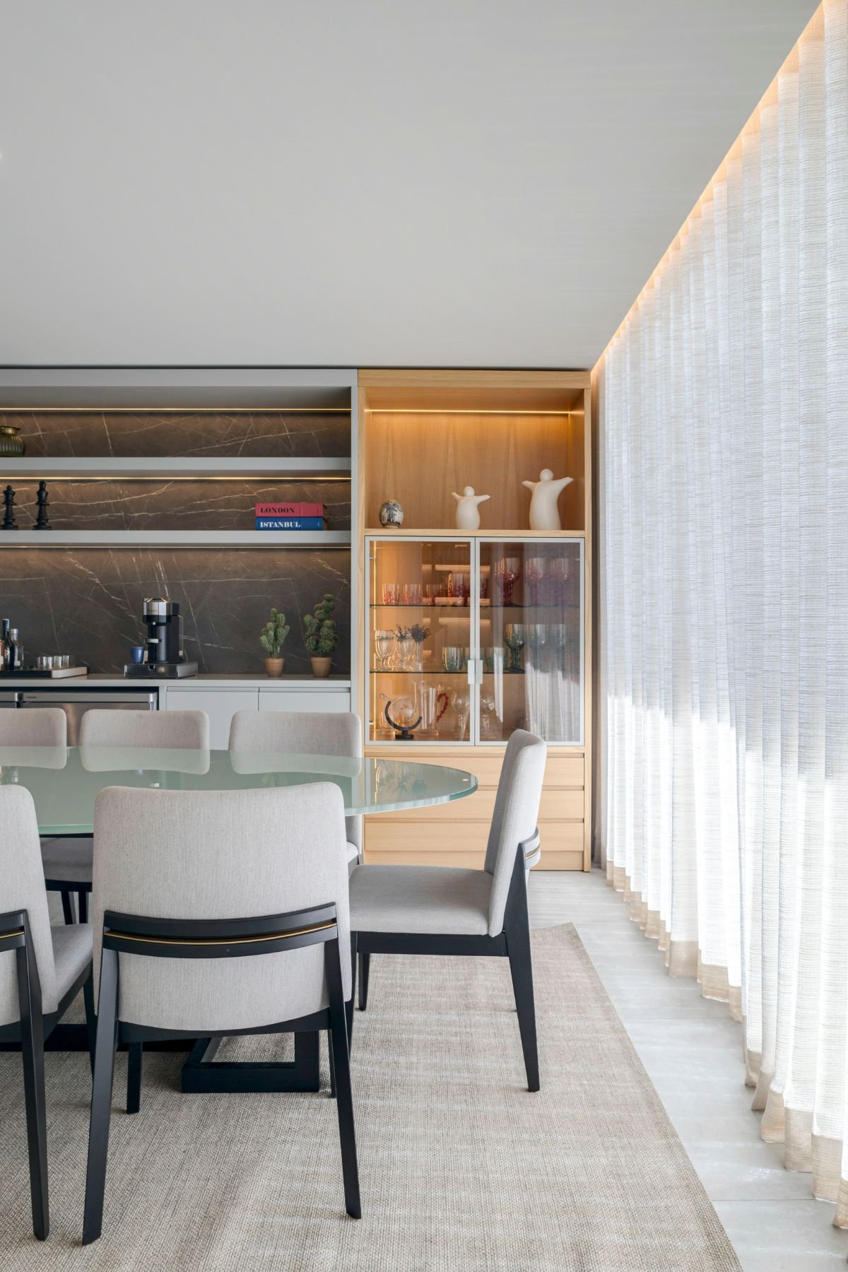 Zona de jantar com mesa de vidro oval e cortinas translúcidas, destacando o móvel bar em madeira e a vitrina com iluminação embutida ao fundo.
