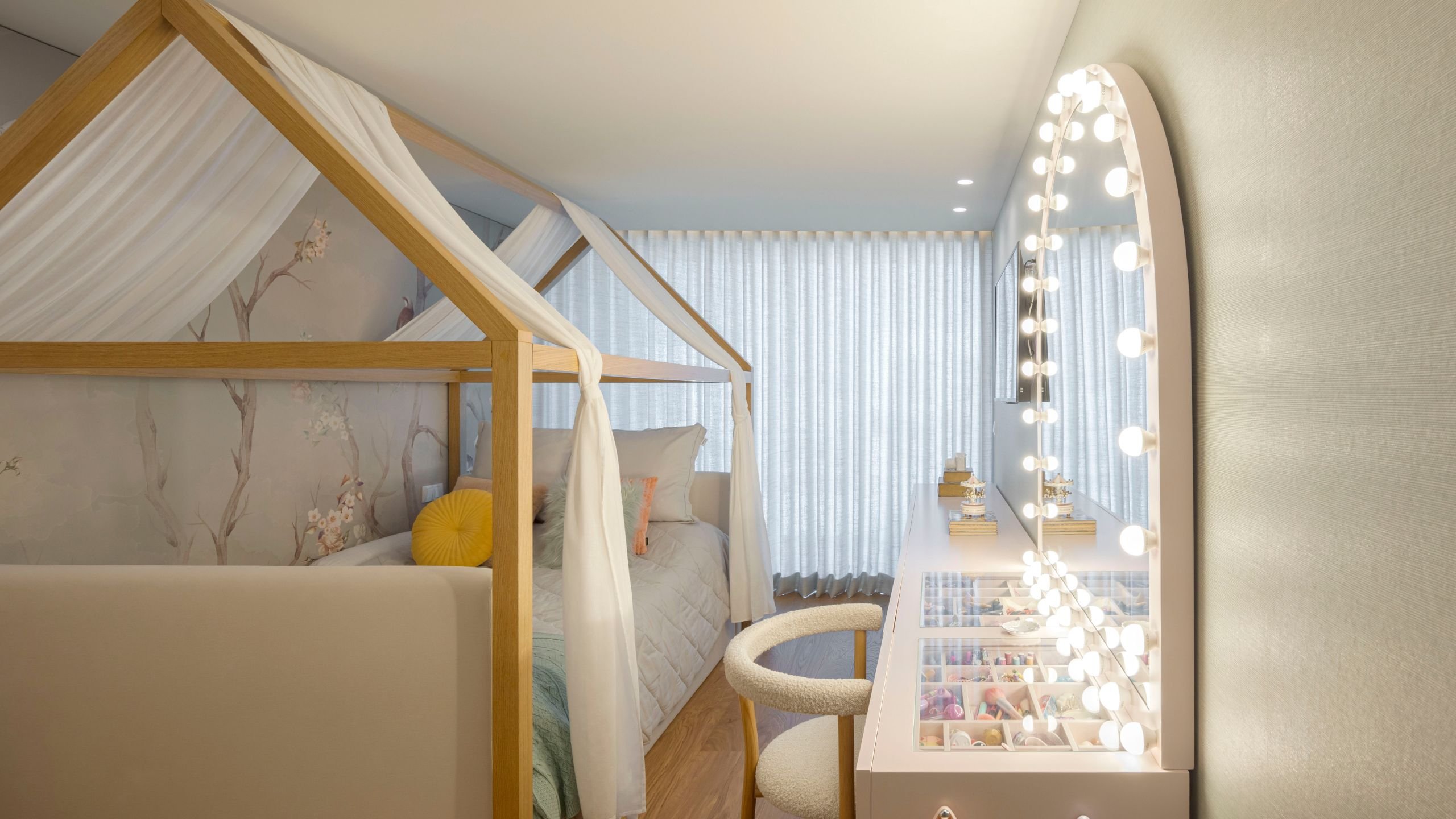 Quarto de criança com cama de dossel em madeira em forma de casa, papel de parede floral suave e toucador com espelho camarim iluminado.
