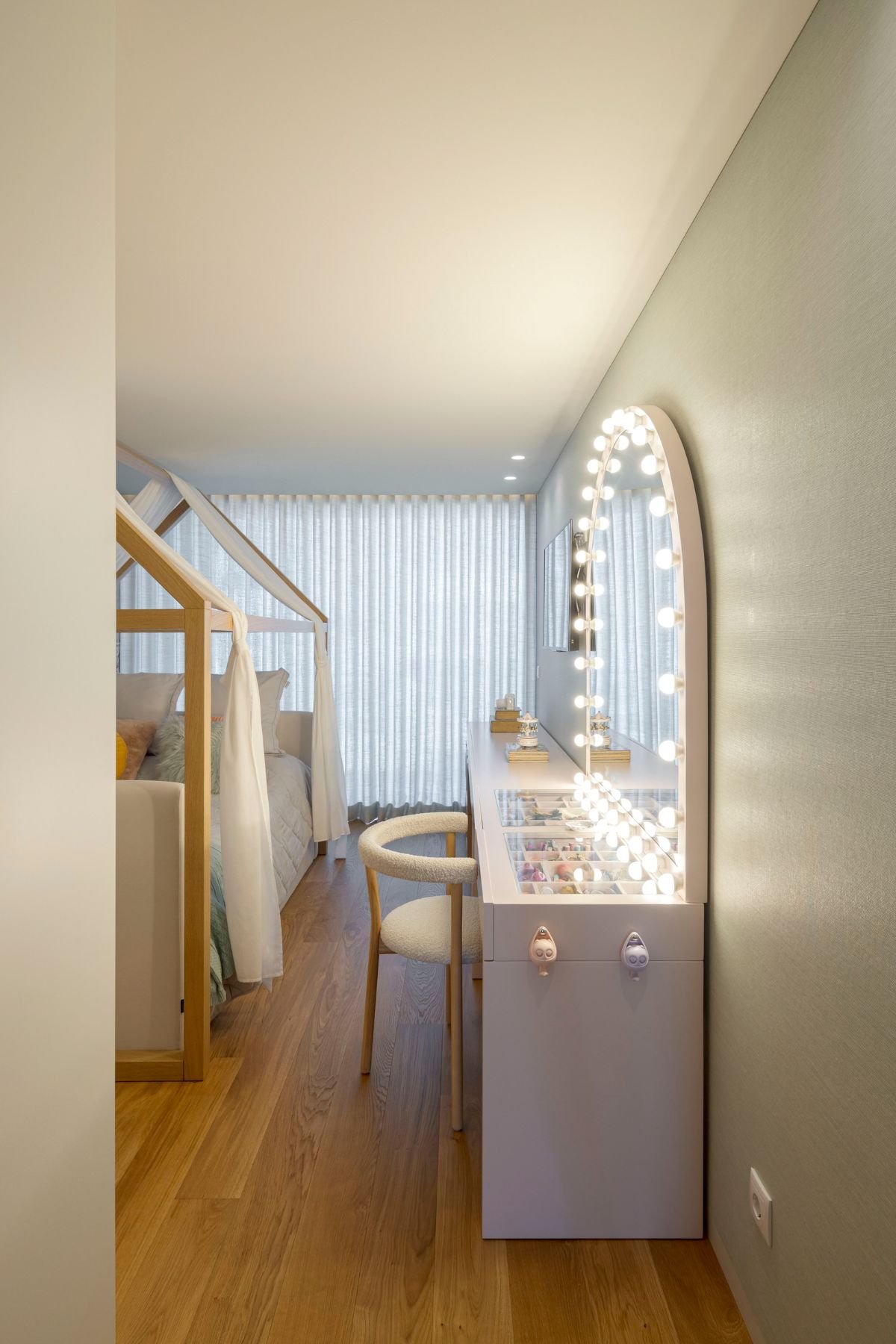 Vista de canto do toucador de maquilhagem branco com tampo em vidro organizador, espelho camarim com luzes LED e cadeira estofada em tecido boucle.