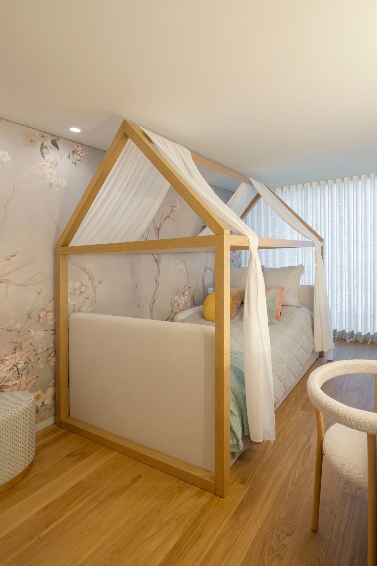 Detalhe da cama infantil com estrutura de dossel em madeira, tecido fluido branco, almofadas decorativas e papel de parede com padrão floral ao fundo.