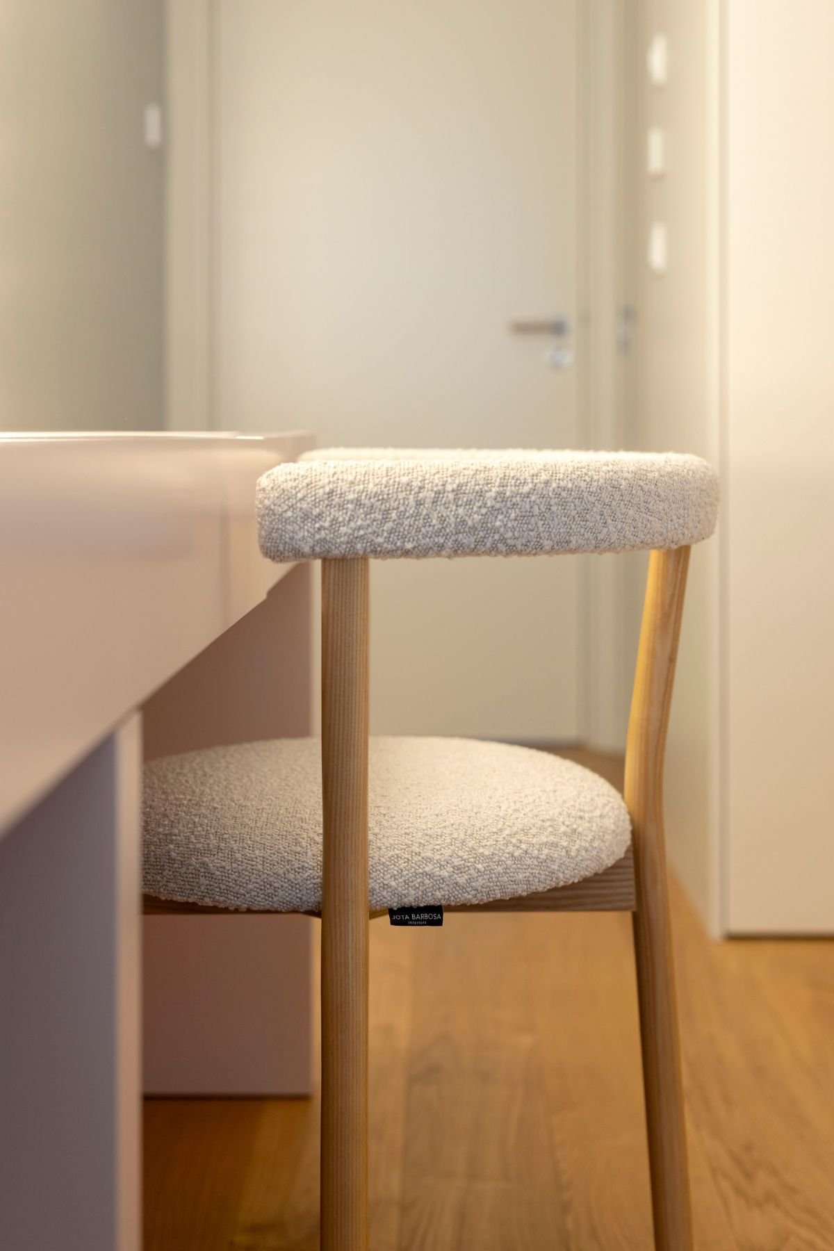 Pormenor do toucador com foco no acabamento da cadeira em madeira com estofo em bouclé claro e etiqueta de design de interiores.
