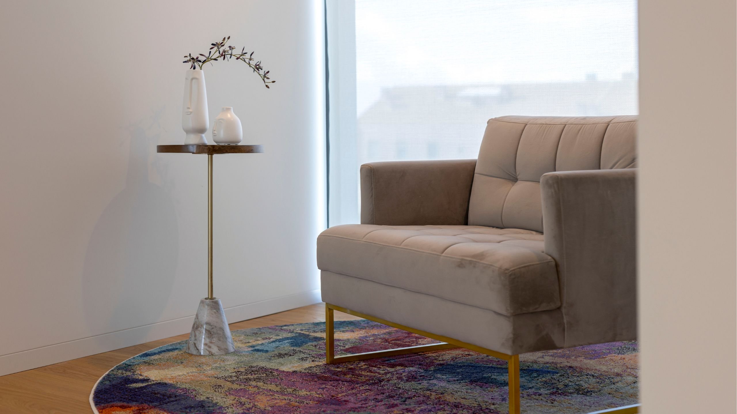 Canto de leitura aconchegante com poltrona estofada bege sobre tapete redondo colorido, acompanhada por mesa de apoio com vaso decorativo.
