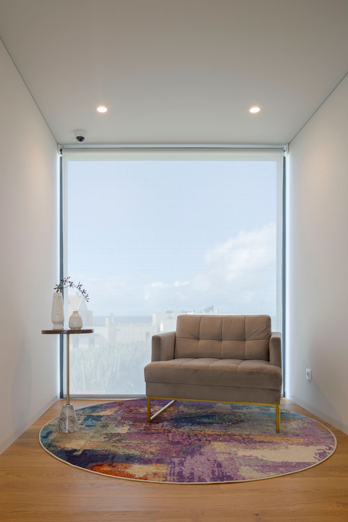 Zona de leitura minimalista junto a uma janela panorâmica, com poltrona confortável em tons neutros, mesa de apoio com base em mármore e tapete circular abstrato.