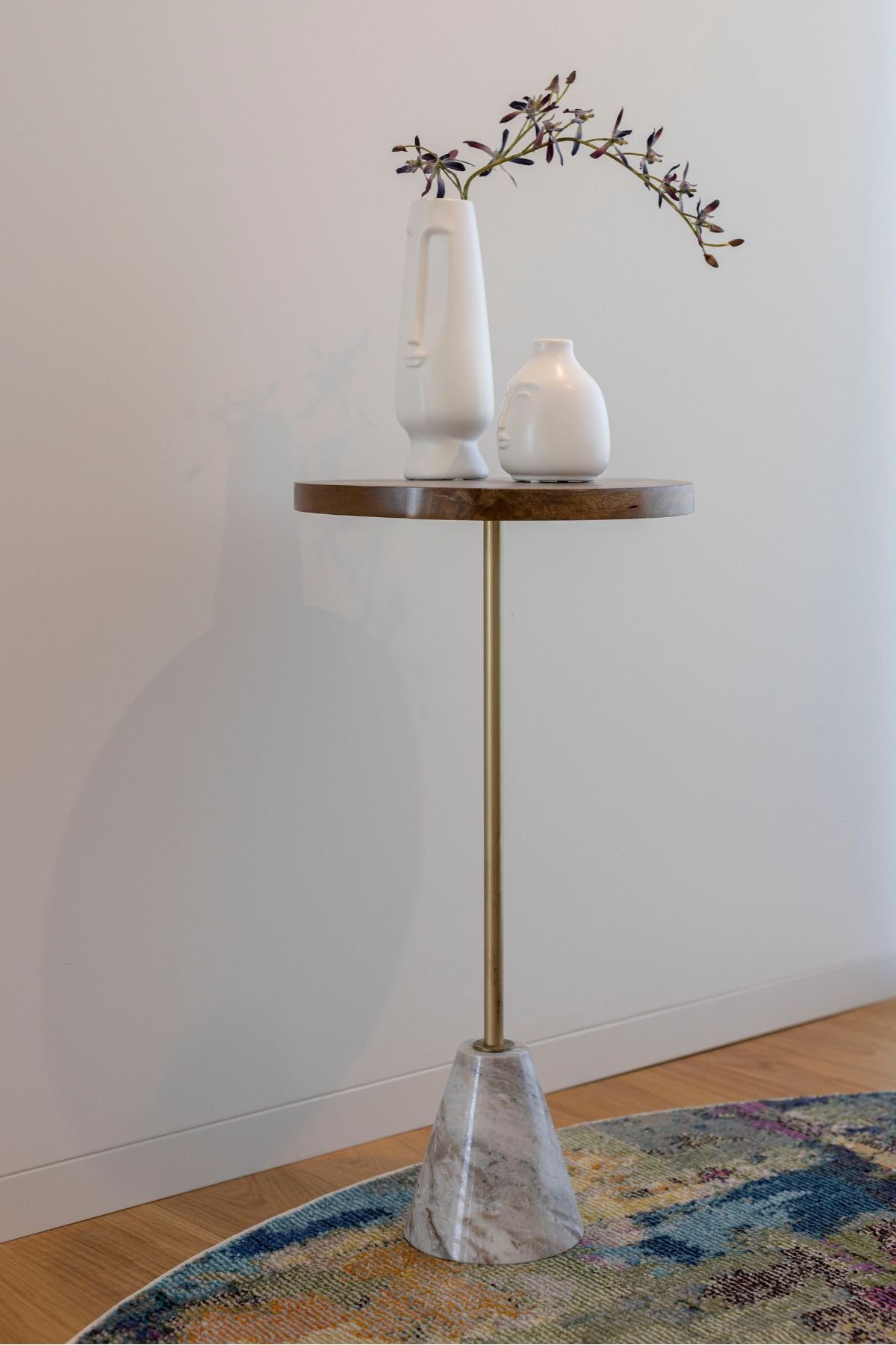 Pormenor de mesa de apoio elegante com base cónica em mármore, haste metálica dourada e tampo em madeira com jarras decorativas brancas e ramo florido.