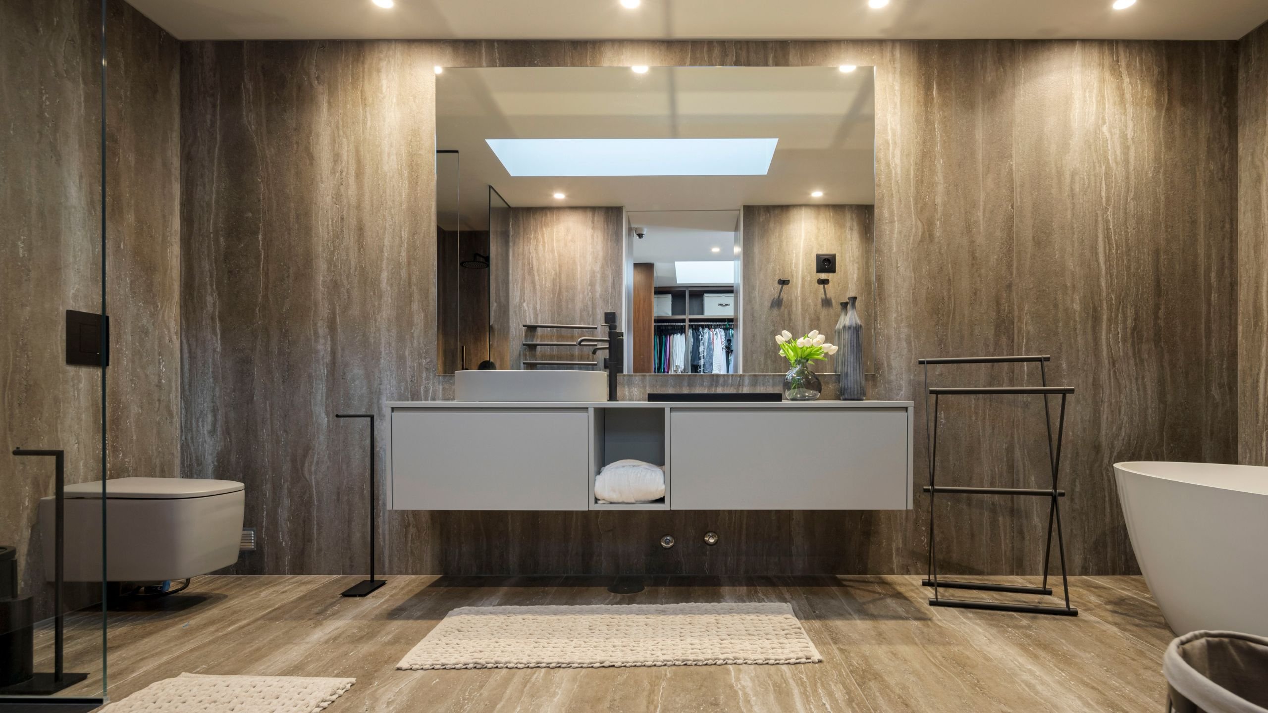 Casa de banho principal de luxo revestida a pedra travertino, com móvel suspenso cinzento, espelho de grandes dimensões, banheira freestanding e torneiras pretas.