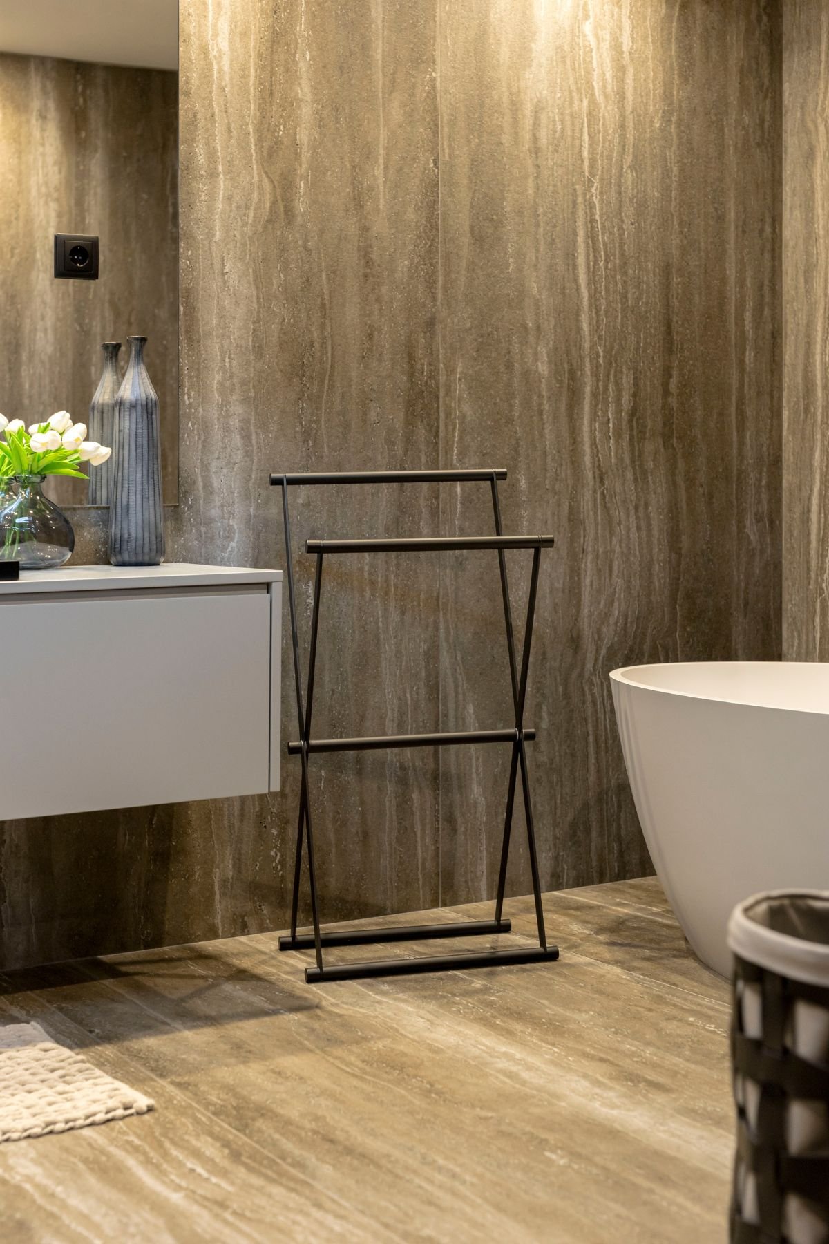 Pormenor do toalheiro de chão em metal preto minimalista ao lado da banheira freestanding e do móvel de lavatório suspenso.