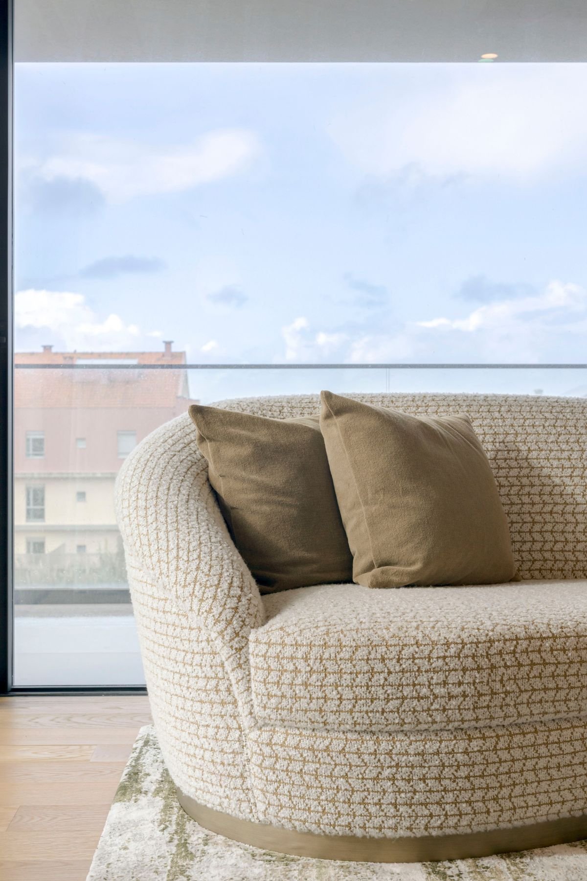 Detalhe do sofá com estofo em tecido padronizado bege e castanho, almofadas decorativas e base em latão discreto junto à janela.