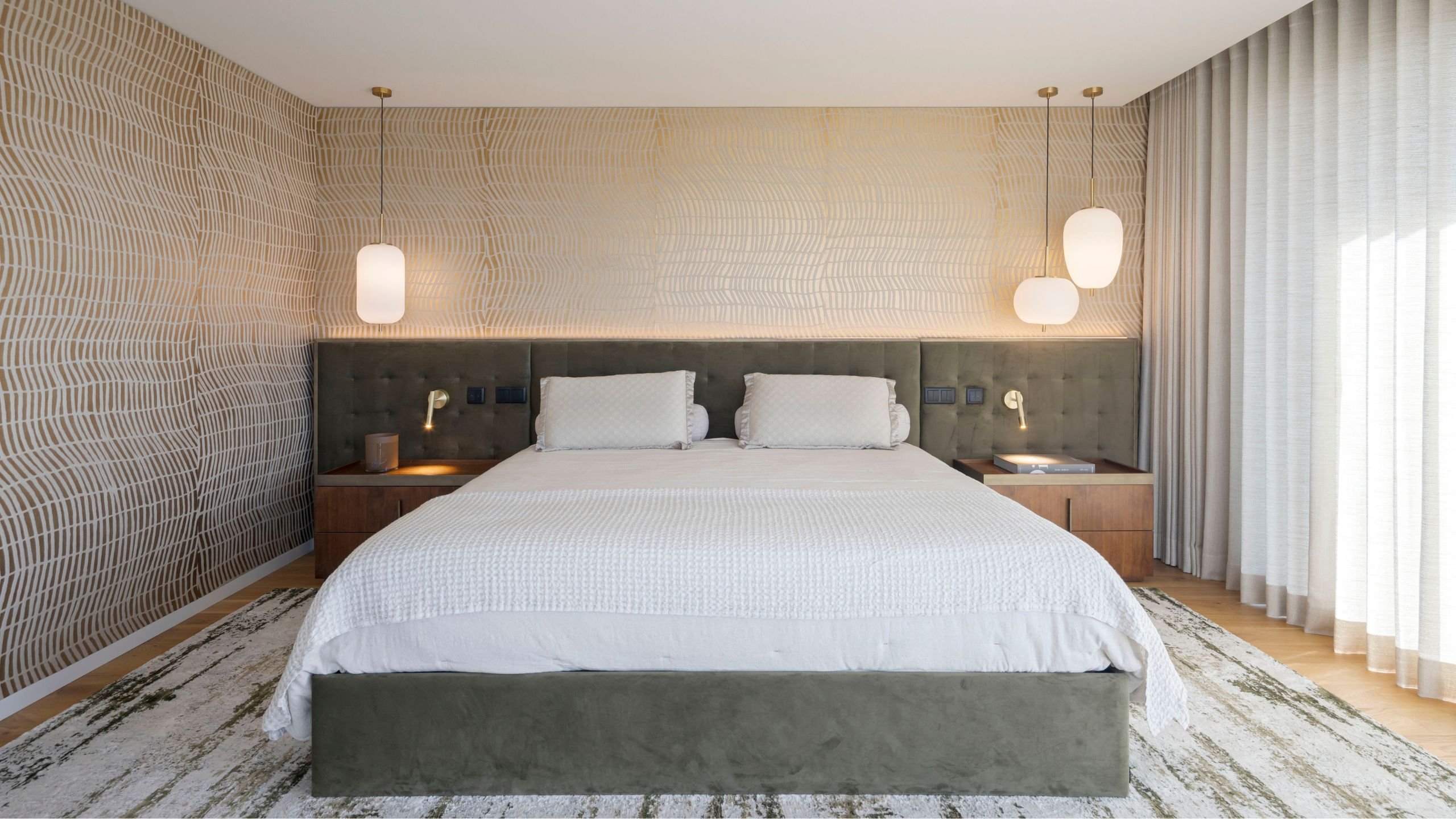 Vista frontal da cama de casal na suíte principal com cabeceira estofada capitonê verde, papel de parede texturizado, candeeiros de suspensão em vidro e tapete abstrato.