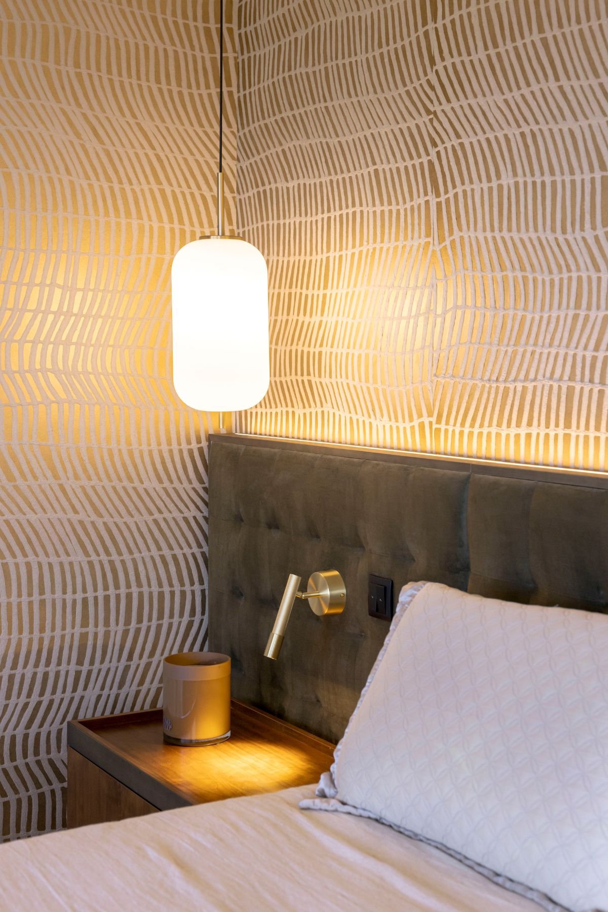 Pormenor do candeeiro de suspensão em vidro branco iluminado sobre a mesa de cabeceira e cabeceira estofada em verde com luz LED embutida.